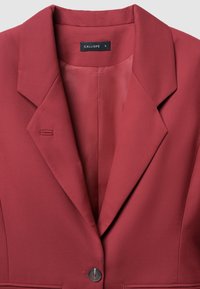 Blazer rosso con risvolti affilati e chiusura a un solo bottone. Tessuto liscio con una sottile texture. Etichetta che riporta "CALLIOPE" e taglia "S."