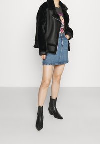 Person som bär svart fuskpäls och läderjacka, grafisk svart t-shirt, blå denimkjol och svarta spetiga ankelskor.