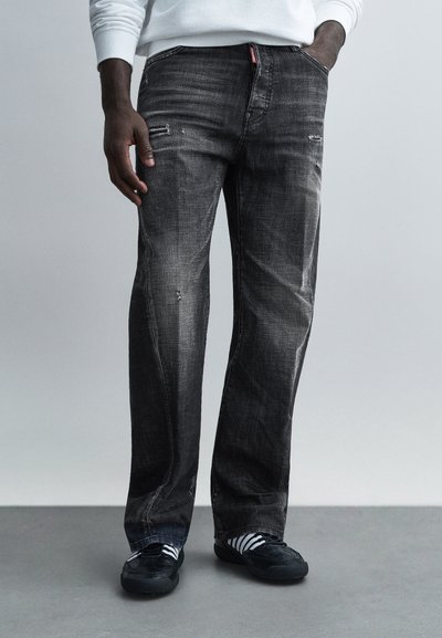 Jean en denim noir à coupe droite. Présente des accents usés, une texture délavée, et des poches avant. Porté avec des baskets noires et des chaussettes rayées.