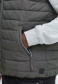 Harmaa puffteliivi, jossa on vaakasuora ommel, erottuva vetoketju ja sivutasku. Varren resorit korostavat teksturoitua kangasta.