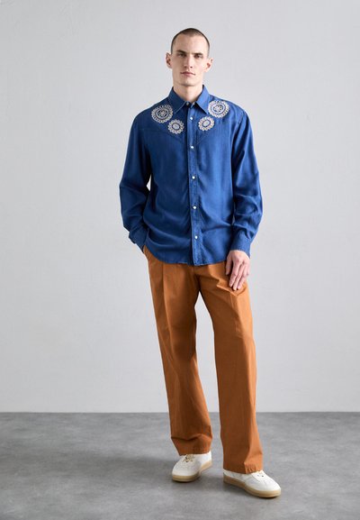Denim-Hemd mit Knöpfen, verziert mit weißen gestickten Blumen auf den Schultern, kombiniert mit orangefarbenen Hosen und weißen Turnschuhen. Glatte Textur.