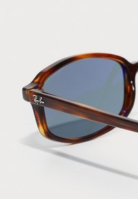 Tortoiseshell Ray-Ban aurinkolasit sinertävillä linsseillä, asetettuna valkoiselle pinnalle, keskittyen vasempaan etuosaan.