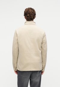 Vyras su trumpais garbanotais plaukais, vilkintis šviesiai smėlio spalvos švarką ir tamsiai pilkas džinsus, stovi priešais baltą fono.