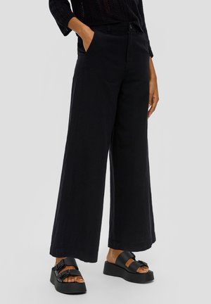 Schwarze Hosen mit weitem Bein aus robustem Stoff, mit Taschen vorne und lockerem Schnitt, kombiniert mit klobigen schwarzen Sandalen.