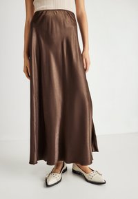 Jupe maxi en satin marron avec une taille élastique, présentant une texture lisse et une légère brillance, associée à des chaussures plates de couleur crème.