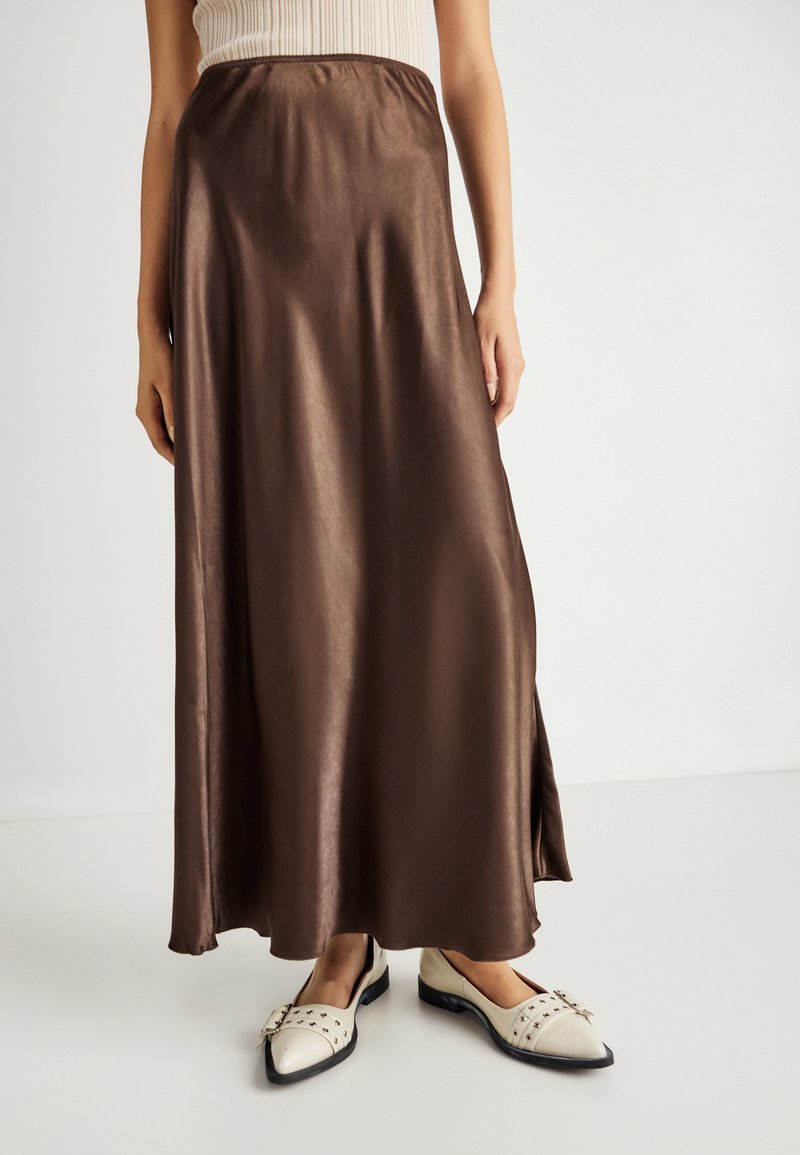 Jupe maxi en satin marron avec une taille élastique, présentant une texture lisse et une légère brillance, associée à des chaussures plates de couleur crème.