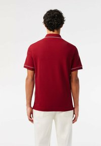 Röd polo t-shirt med korta ärmar, ribbad krage och vit accentdetalj. Bomullstyg med en slät textur. Sedd bakifrån.