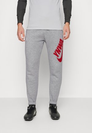 Hombre vistiendo pantalones deportivos grises con el logo rojo de Nike en la pierna derecha, zapatillas negras y una camiseta de manga larga blanca y negra, de pie sobre un fondo liso.