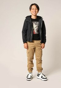 Veste noire à zip avec capuche à cordon, t-shirt graphique noir, pantalon cargo en kaki et baskets noir et blanc avec lacets.