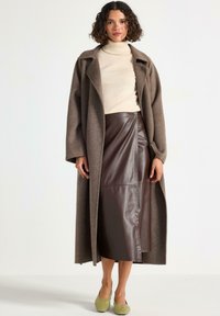Lang brun uldblandingsfrakke med en bred krave, lagdelt over en beige turtleneck og en brun læder midi-nederdel, parret med grønne flade sko.
