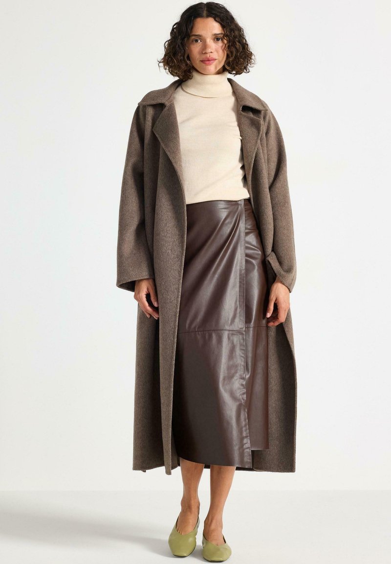 Lang brun uldblandingsfrakke med en bred krave, lagdelt over en beige turtleneck og en brun læder midi-nederdel, parret med grønne flade sko.