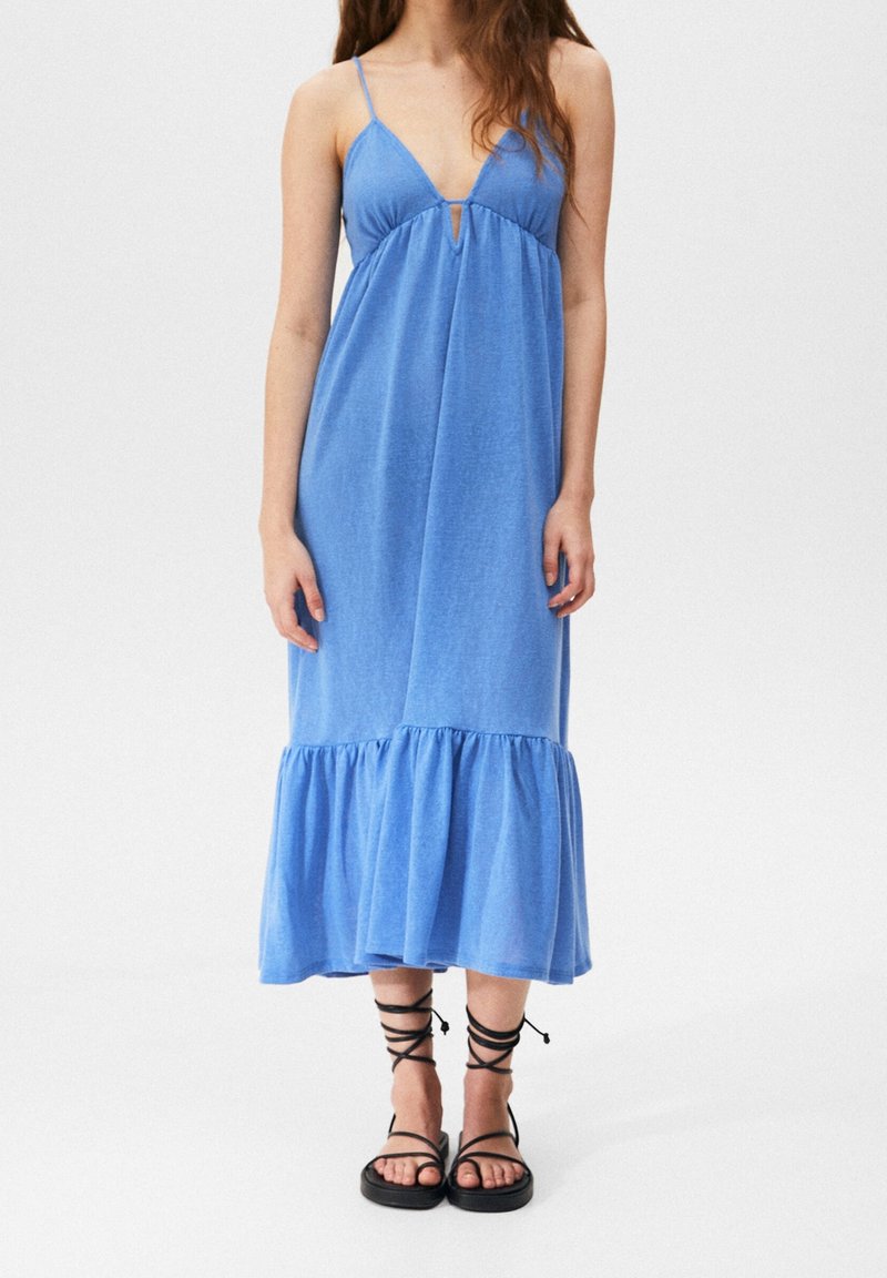 Femme portant une robe midi bleue sans manches avec de fines bretelles et un ourlet à volants, associée à des sandales plates noires à lacets sur fond blanc.