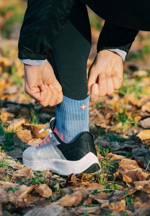Person pulling up a blue sock with a small flag emblem while wearing black leggings and white running shoes outdoors on fallen leaves.