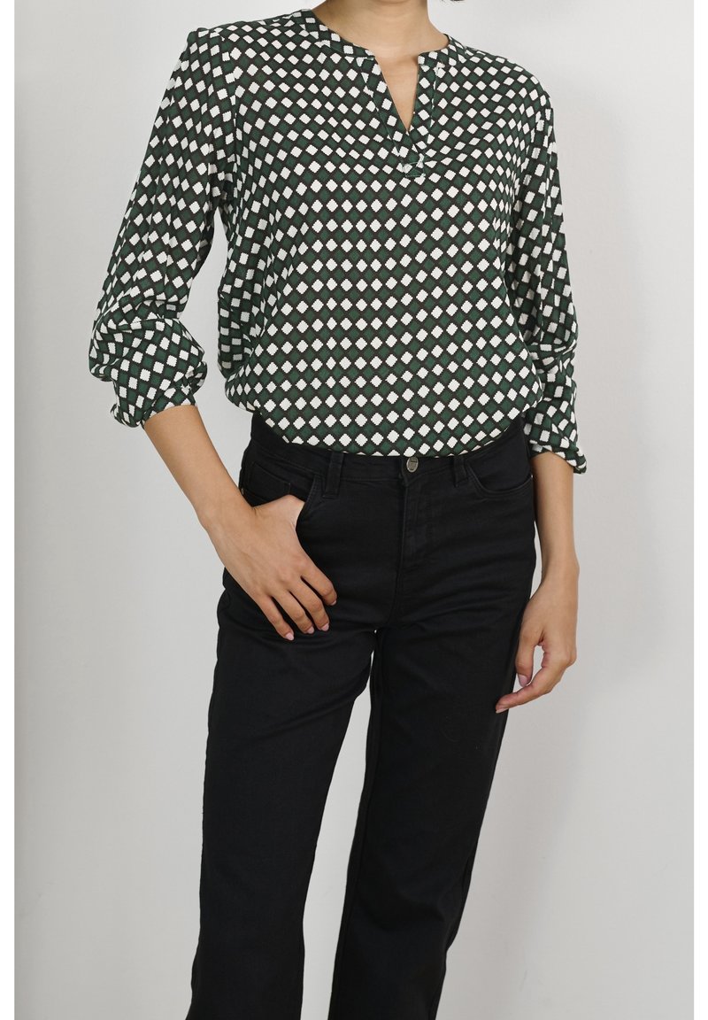 Blouse verte à motifs avec des formes en losange blanches, dotée d'un col en V et de manches retroussées, associée à un jean noir. Texture de tissu lisse.