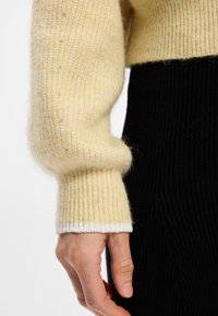 Pull en tricot de couleur jaune pâle avec une texture côtelée et un accent blanc au niveau des poignets, porté sur un vêtement noir texturé, mettant en valeur un détail de la main.