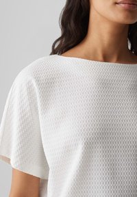 Hvid tekstureret bluse med et vævet mønster, bred halsudskæring og korte ærmer, der fremhæver et blødt og let stof.