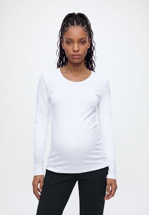 Jeune femme enceinte aux cheveux tressés, portant une chemise blanche à manches longues et un pantalon noir, debout devant un fond blanc uni.