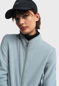 Svijetloplava flis jakna s visokim ovratnikom i patentnim zatvaračem, s teksturiranim površinom. Nosena preko crnog rolka. Crna kapa na glavi.