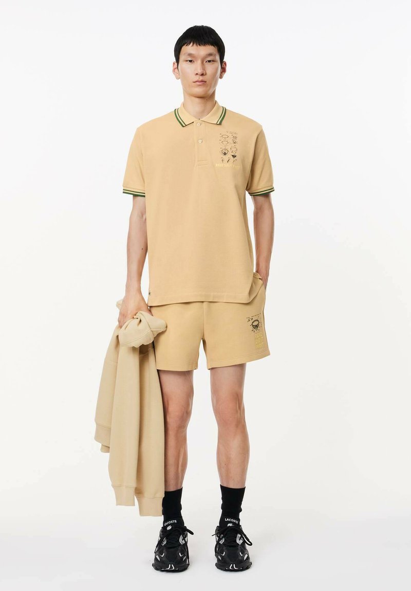 Camisa polo color tostado con rayas verdes, con un diseño gráfico en el pecho. Combinada con pantalones cortos a juego y un sudadera ligera colgada del brazo.