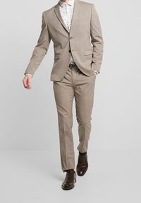 Traje marrón claro con corte ajustado, tejido texturizado, solapas en pico y dos bolsillos frontales. Combinado con una camisa de estampado y zapatos de vestir marrones.