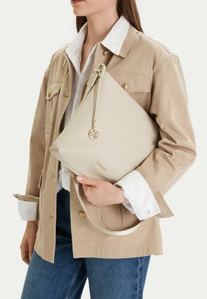 Persona che indossa una giacca beige e una camicia bianca che tiene una borsa a tracolla in pelle color crema con logo dorato e ciondolo rotondo.