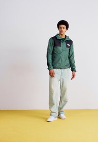 Grüne Windjacke mit schwarzen Akzenten, Knopfverschluss, zwei Brusttaschen, kombiniert mit hellblauen Jeans und weißen Sneakern vor einem gelben Hintergrund.