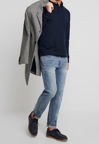 Muž oblečený v světle modrých džínech, námořnickém propínacím tričku s dlouhým rukávem, námořnických semišových botách a nese přes rameno kabát s houndstooth vzorem v černobílé barvě.