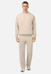 Bežs sporta krekls un atbilstoši bikses no mīksta, teksturēta materiāla, ar brīvu piegriezumu un ribotām aprocēm, valkāts kopā ar gaišiem sneakers.