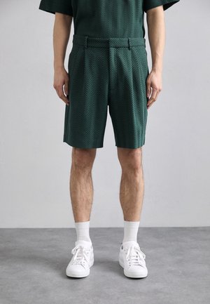 Green shorts in a herringbone pattern, waist pleats, side pockets, paired with white sneakers and socks, suitable for casual wear.