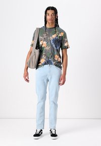 Mørk t-skjorte med blomsterprint, lyse blå jeans, svart-hvite sneakers og en grå budveske. Modellen står med hendene på sidene.