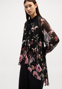 Blouse noire transparente avec des motifs floraux en rose et blanc ; dotée d'un col, de manches longues et d'un design fluide avec un ourlet asymétrique.