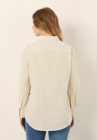 Camisa de manga larga color crema en lino ligero con cuello puntiagudo, yugo trasero y puños abotonados. Presenta un dobladillo curvado.