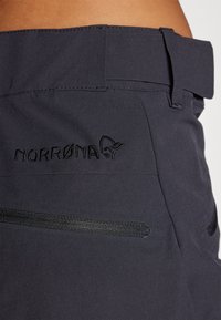 Dark fabric pant featuring a wide waistband, Norrøna logo embroidered in black, and two textured accent strips on the front.