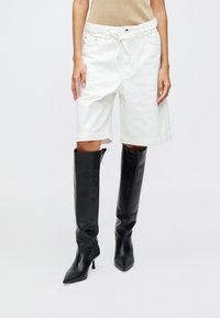 Valkoiset farkkuhousut, joissa on rento malli ja naru vyötäröllä, yhdistettynä korkeisiin, kiiltäviin mustiin nahkasaappaisiin.