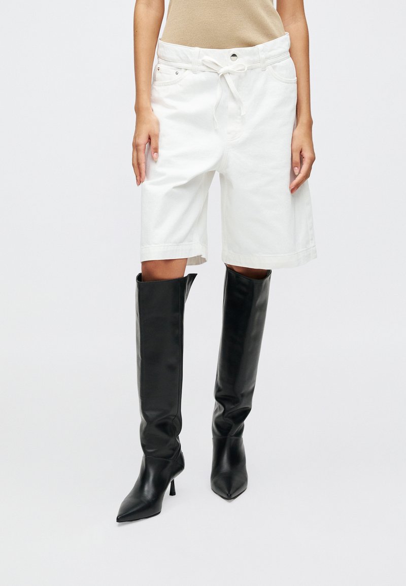 Valkoiset farkkuhousut, joissa on rento malli ja naru vyötäröllä, yhdistettynä korkeisiin, kiiltäviin mustiin nahkasaappaisiin.