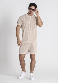 Beige gebreide shirt met korte mouwen en een knoopsluiting, gecombineerd met bijpassende shorts. Witte sneakers en donkere zonnebrillen maken de look compleet.