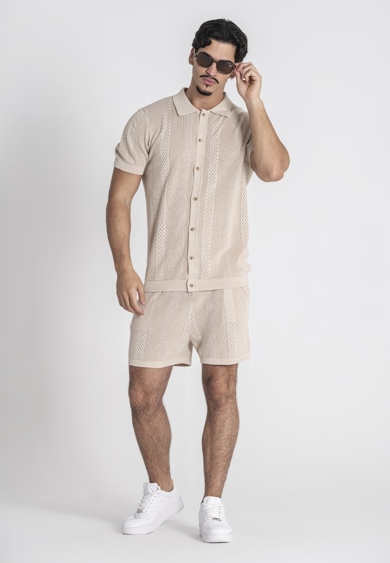 Beige gebreide shirt met korte mouwen en een knoopsluiting, gecombineerd met bijpassende shorts. Witte sneakers en donkere zonnebrillen maken de look compleet.