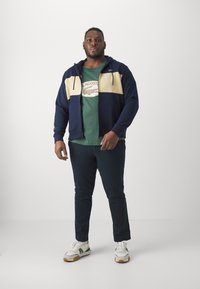 Hoodie v námorníckej a krémovej farbe s rozopínaním, zelené tričko s logom, tmavé nohavice a biele tenisky so zelenými akcentmi.