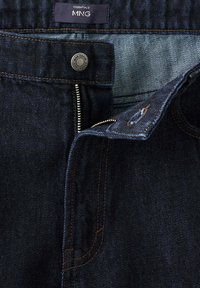 Jean en denim bleu foncé avec fermeture éclair partiellement ouverte, fermeture par bouton et étiquette de marque MNG Essentials à l'intérieur de la ceinture.