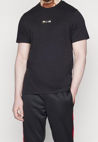 Musta puuvillane T-särk, millel on ümmargune kaelus ja kuldne "AIR" logo rindkere piirkonnas. Kombineeritud mustade pükstega, millel on punased külgmised triibud.