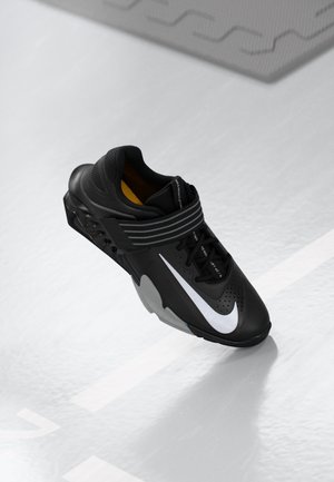 Scarpa sportiva nera con il logo Nike bianco, dettagli grigi e una spessa fascia a righe sopra i lacci neri, esposta su una superficie chiara.