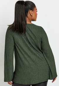 Top lungo a maniche lunghe di un verde scuro strutturato, con un ampio scollo e maniche svasate. Il tessuto presenta un motivo diagonale sottile, che aggiunge profondità.