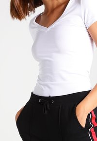 Mujer vistiendo una camiseta blanca ajustada con cuello en V y pantalones negros con cordón, con franjas laterales rojas y blancas, con las manos en los bolsillos frente a un fondo blanco.