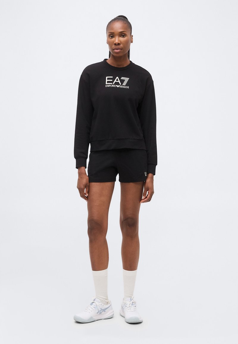 Mujer de pie con sudadera negra y pantalones cortos EA7 Emporio Armani, calcetines blancos y zapatillas deportivas blancas sobre fondo blanco.