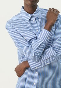 Blue and white vertically striped button-up shirt with a collar, featuring two front pockets and buttoned cuffs. Smooth fabric texture.