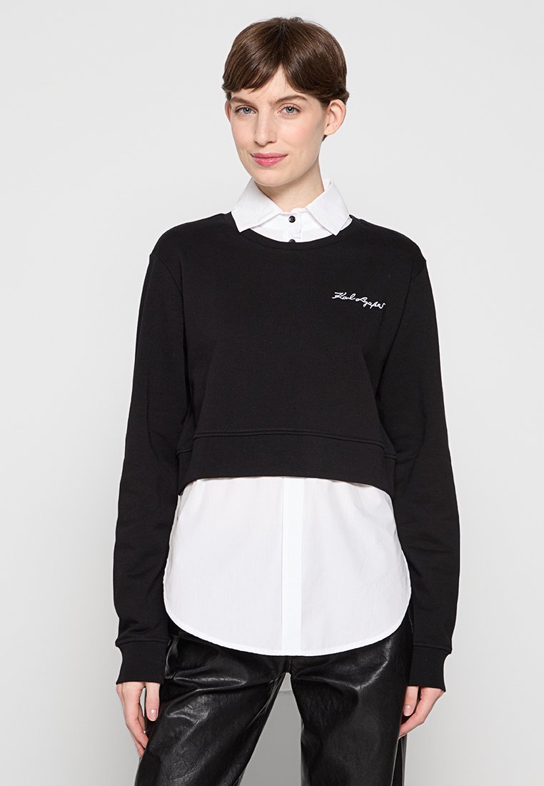 Sweatshirt noir court avec des poignets côtelés, comportant une chemise blanche en dessous. Texte brodé blanc à l'avant du sweatshirt.
