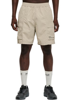 Mand iført beige cargoshorts med sidelommer, hvide logo-strømper og sorte sneakers, stående mod en hvid baggrund.