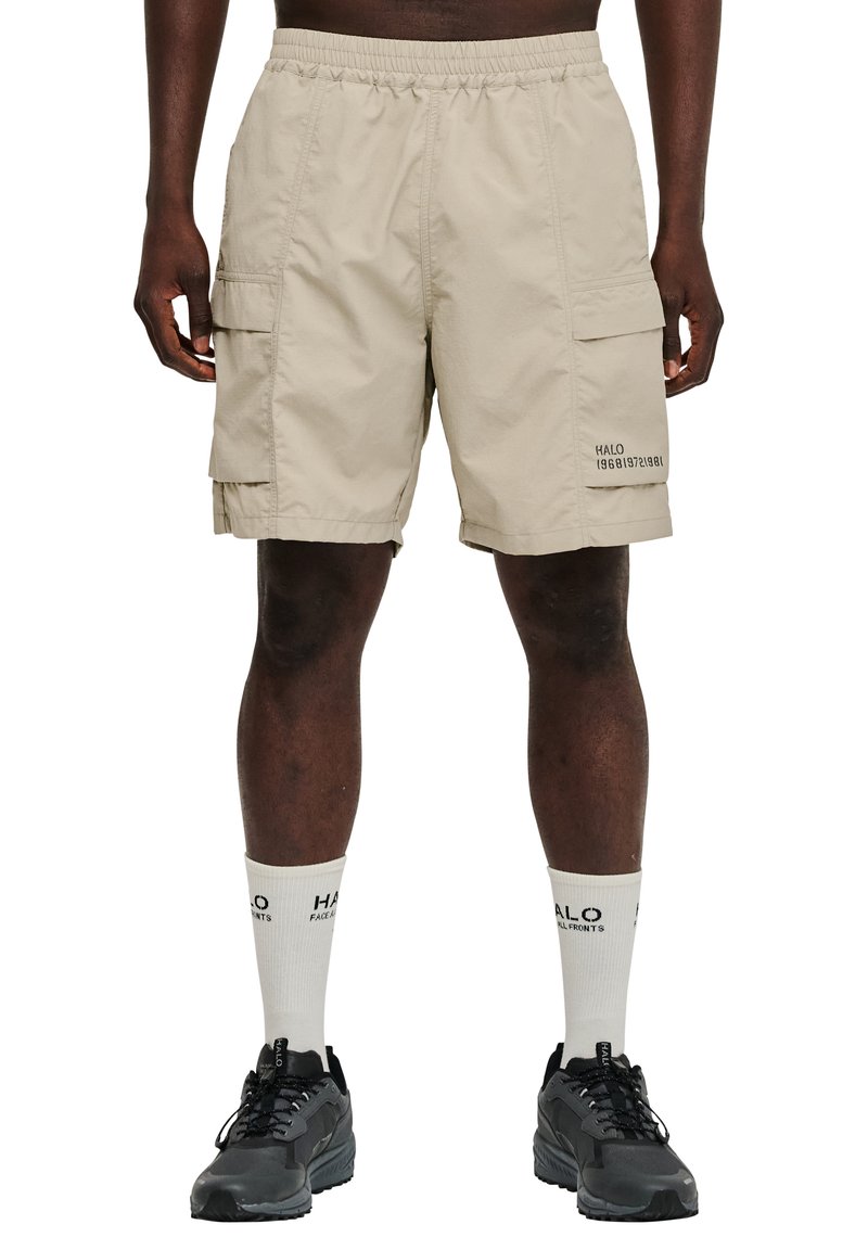 Man draagt beige cargoshorts met zijzakken, witte sokken met logo en zwarte sneakers, staand tegen een witte achtergrond.