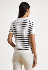 Sieviete ar gariem tumšiem matiem, tērpusies īsroku baltā džemperī ar tumši ziliem horizontāliem svītrām un gaišām biksēm, skatās prom no kameras.