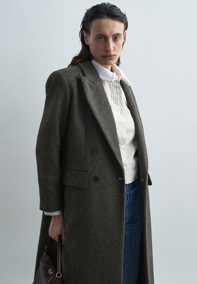 Dark green double-breasted coat with lapels, featuring black buttons, worn over a light plaid shirt and denim, held with a brown handbag.